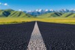 © oksa_studio - Low angle view of a wide asphalt road with white dividing lines leading towards a blurred background of green hills and snowy mountains under a blue sky