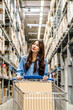 © TeTe Song - Young asian woman navigates through a vibrant home goods store in warehouse, pushing her shopping cart as she scans the shelves filled with a variety of stylish products, searching for inspiration