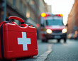 © Vadym - Red first aid kit stands on city street. Ambulance car with flashing lights drives, urgent response. Focus on kit with medical cross symbol, emergency medical service.