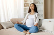 © Anastasiya - Beautiful young pregnant woman practicing yoga at home, sitting in lotus position on couch in living room, expectant female meditating with closed eyes, making meditation for wellness