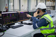 © JD Studio - Technician in safety helmet uses computer systems to control and monitor substation operations with multiple screens displaying data and control panels in industrial setting