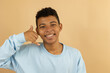 © Answer 7 - Cheerful brown skinned Latino teenager in a pastel blue sweatshirt making a call gesture with his hand on a light brown background with copy space, concept of communication, connection