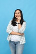© ArvStd - Portrait of cheerful young asian woman, smiling looking at camera, folding hands, touching cheek with finger, standing isolated on blue background.