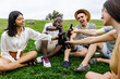 © Xavier Lorenzo - Group of young friends enjoying a summer day, sitting on grass, clinking bottles, and sharing laughter in a relaxed outdoor setting