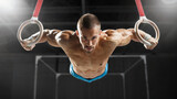 Male gymnast performing a strength move on gymnastic rings in a dark gym. Intense expression, muscular body, dramatic spotlighting. Front angle with black background.