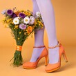 © MOLNAR - A pair of orange high-heeled shoes with ankle straps, worn by a person with lavender tights. A colorful bouquet of flowers is held in front against a yellow background.