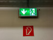 © Kateryna Muzhevska - Emergency exit sign with illuminated green arrow and running figure, positioned above fire extinguisher symbol on wall, indicating safety direction