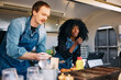 © Maskot - Curly haired woman taking order while male coworker making hot dogs in food truck