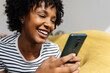 © Xavier Lorenzo - Happy young Afro American girl having fun using mobile phone relaxing on sofa at home