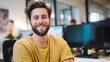 © Jittapon - A young happy and confident man with a beard and curly hair sitting at his desk in a modern office environment surrounded by technology and communication equipment
