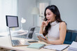 © Tj - Focused young woman in an office setting, multitasking with a smartphone and laptop. She appears engaged in her work, surrounded by notebooks and a modern workspace environment