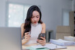 © Tj - Focused businesswoman using her smartphone while working at a desk, surrounded by documents and a laptop, in a modern office environment with soft natural light