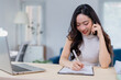 © Tj - Young professional woman efficiently multitasking at her desk, talking on the phone while taking notes. The setting is a modern office with a laptop and bright, airy atmosphere