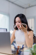 © Tj - Professional woman in a modern office using a smartphone for communication while working on a laptop. Bright, focused environment with a sense of productivity and efficiency