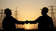 © Qrryferry - Two silhouetted workers shaking hands at sunset with power lines and towers in background