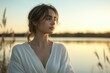 © hkjdesign - A young woman in a white dress gazes thoughtfully at a tranquil sunset over a lake.