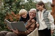 © Seventyfour - Senior Caucasian man sitting outdoors in village holding large book while smiling with two children, boy and girl, standing close and looking at book together