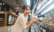 © dodotone - Young asian woman customer supermarket shopping cart choosing dairy products beverages shelf grocery food at department store