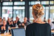 © amaf - A blonde woman with her hair in a bun, seen from behind, presents to a blurred audience in a modern, bright room, a laptop visible before her