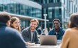 © Gamenin - Diverse group of young adults smiling, engaged in conversation outdoors at a table with a laptop, amidst a blurred cityscape backdrop