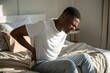 © BlackBird Studio - Young African American Man Dealing with Back Pain in Bedroom During Morning Routine