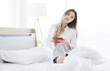 © Bangkok Click Studio - Asian young happy beautiful woman relaxes, enjoys a peaceful moment sitting holding using digital tablet computer under white blanket on bed in cozy bedroom bright weekend lifestyle morning at home.