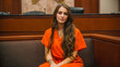 © MACKY - a woman in an orange jumpsuit sits in a courtroom setting. The woman has long brown hair and is wearing jewelry. She is sitting in a wooden booth and appears to be waiting