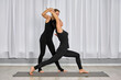 © TRAVELARIUM - Two adult women practice yoga in serene studio. Instructor assists student in warrior pose. Bright lighting and minimalist design create calmness