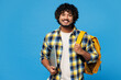 © ViDi Studio - Young smiling smart IT Indian boy student wear yellow casual clothes backpack bag hold closed laptop pc computer look camera isolated on plain blue background. High school university college concept.