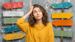 © Naufal - A young woman standing in front of multiple signposts, looking confused and uncertain, searching for direction, outdoors, decision making, dilemma, travel and guidance concept.