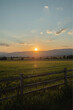 © MDZAHANGIR - The sunset scene in a serene countryside features a warm golden glow, a wooden fence, a lush green and yellow field, and soft clouds adding depth to the serene scene.