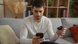 © Krakenimages.com - Handsome young man sitting indoors using smartphone holding credit card in home living room setting suggesting online shopping or mobile banking