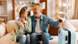 © Home-stock - Joyful retired senior couple ready for trip, sitting on sofa at home, looking and smiling at each other, waiting for vacation