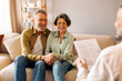 © Home-stock - Happy European senior spouses holding hands while having consultation with real estate agent or psychologist, sitting on sofa. Specialist's service offer