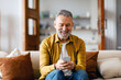 © Home-stock - Senior man using smartphone texting, networking in social media or scrolling social media, sitting on sofa at home. Gadgets and retirement lifestyle