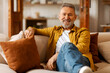 © Home-stock - Contented handsome senior man sitting on sofa and smiling at camera, retired male relaxing in living room. Retirement lifestyle, domestic comfort concept