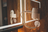 Wall-mounted white hotel hair dryer with coiled cord, reflected in illuminated bathroom mirror. Modern bathroom interior with wooden panels and warm lighting