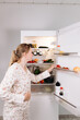 © Kateryna Muzhevska - Woman in floral pajamas stands by open refrigerator, reaching for fresh fruits and vegetables, showcasing a healthy lifestyle and the importance of nutrition in daily meal preparation