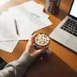 © ma--photo - Cozy top view of a hand cradling a mug of hot chocolate topped with marshmallows, set on a warm-toned work desk with papers and soft light. Suggests comfort during a busy workday copy
