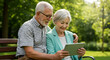 © mumtaz - Senior couple enjoying time outdoors using a tablet device, sitting on a park bench with lush green trees and sunlight in the background for a relaxing, happy atmosphere