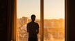 © A R - Person stands by bright window overlooking buildings, looking at a sunrise or sunset, contemplating future goals.
