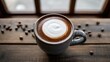 © Xiaotong - Hot cappuccino with simple latte art in a ceramic mug on a rustic wooden table with scattered coffee beans by a window.