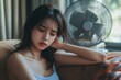 © Snap Mania - Suffering from a summer heatwave stroke, tired Asian young woman sitting on the couch in the living room without air-conditioning, using a fan for ventilation, Generative AI
