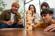 © Chanakon - Young Asian friends are happily playing wooden block together at home, enjoying their leisure time. Group friends carefully remove a wooden block while others watch, having fun a classic table game.