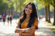 © Pete - Joyful young Mexican woman with long black hair smiles broadly, arms crossed outdoors on a sunny day. She radiates happiness and confidence. Background shows blurred park path with trees and people.