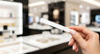 © Aiustock - Woman's hand holding a white perfume test strip in a blurred luxury boutique, sampling fragrances and cosmetics. Focus on scent testing in store.