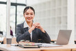 © kenchiro168 - Confident businesswoman in gray suit sitting at desk with laptop, coffee cup, and documents in modern office, smiling and looking at camera, professional and approachable atmosphere