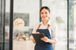 © kenchiro168 - Young Asian woman wearing apron standing at cafe entrance holding digital tablet, smiling confidently, modern small business owner, welcoming atmosphere, bright interior