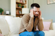 © Dorde - Young Caucasian Man Sitting on Sofa Holding Head in Pain in a Comfortable Living Room Setting
