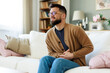 © Dorde - Young Man in Casual Outfit Sitting on Couch with Stomach Pain in Bright Comfortable Living Room